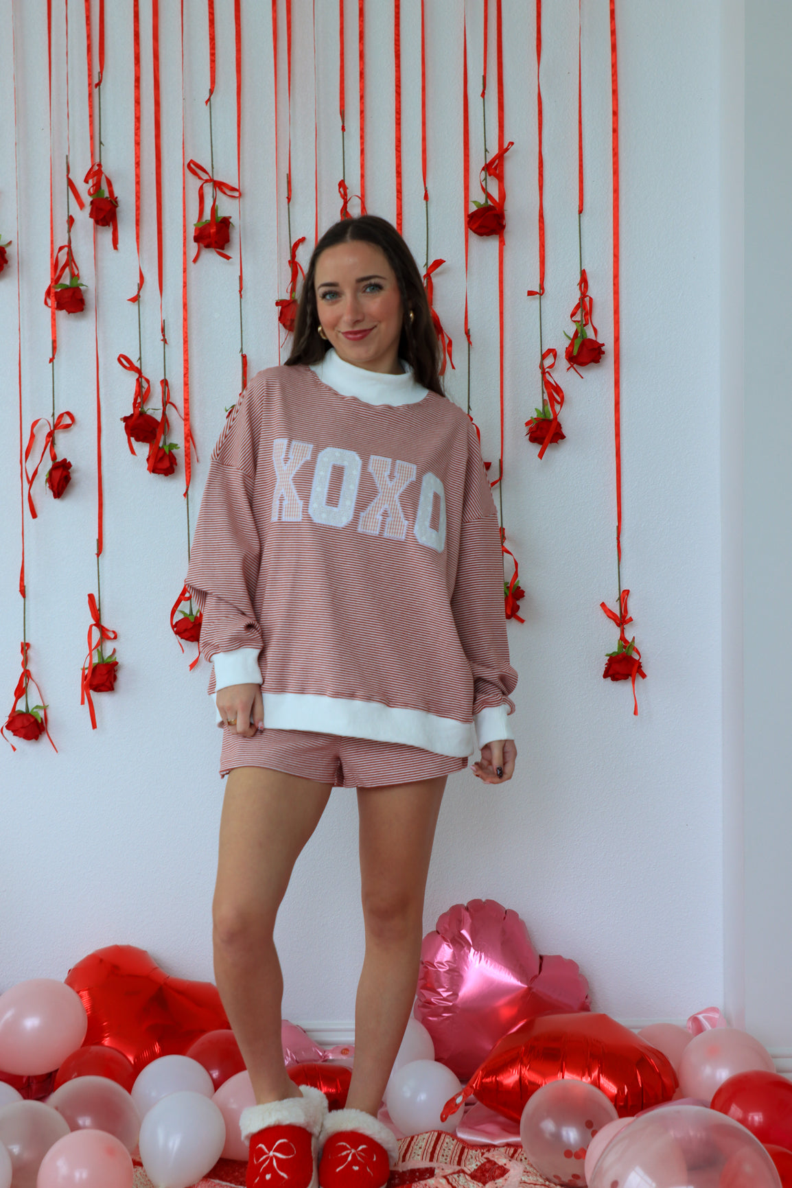 Woman in a pink 'XOXO' sweatshirt and shorts standing in front of a decorative wall with red ribbons and balloons.