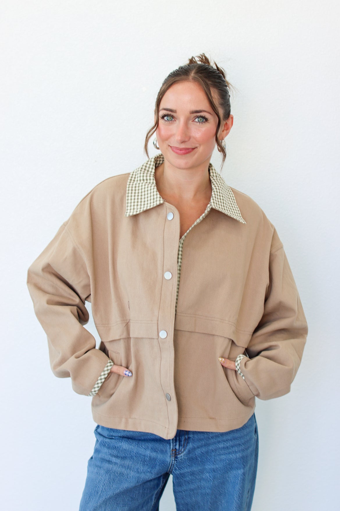 A woman wearing a beige utility jacket with a green and white gingham patterned collar, paired with blue jeans, standing against a plain white wall.