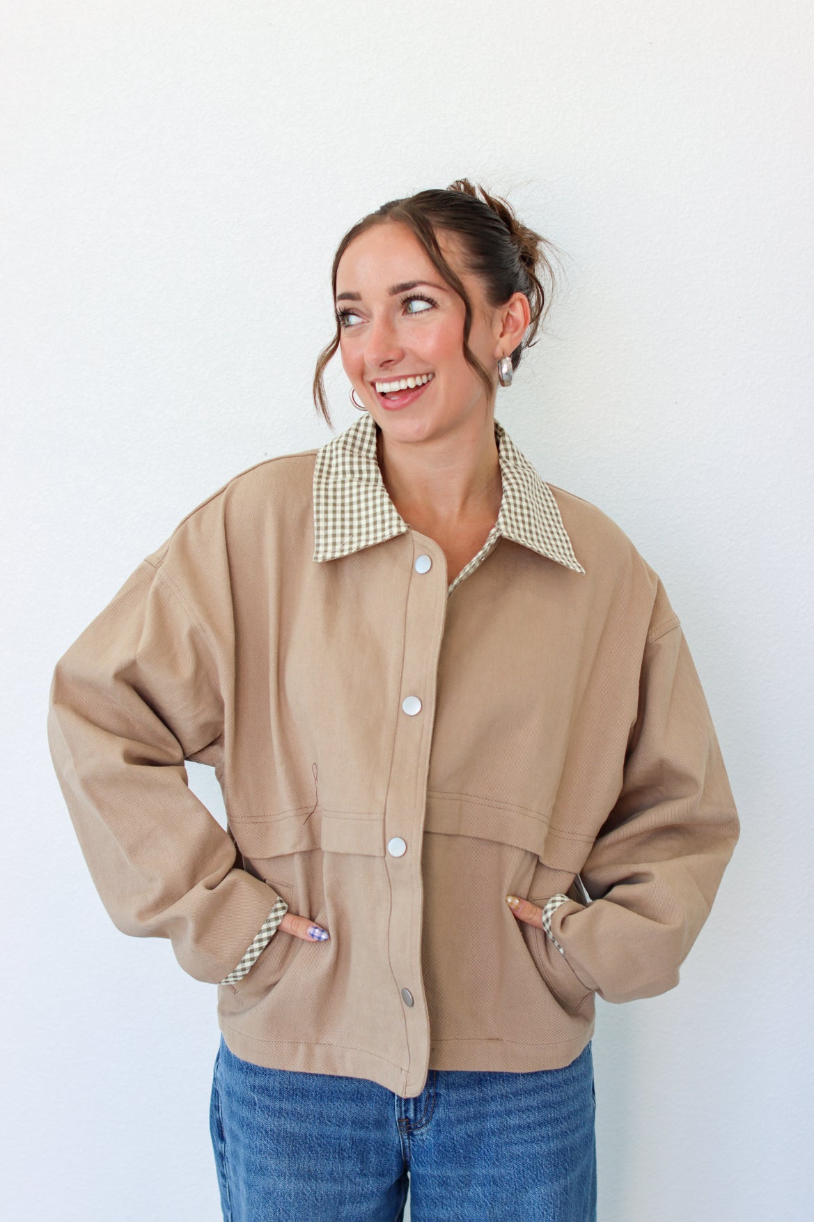 A woman wearing a beige utility jacket with a green and white gingham patterned collar, paired with blue jeans, standing against a plain white wall.