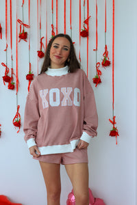 Woman wearing a pink 'XOXO' sweatshirt and shorts in front of a decorative wall with red ribbons and flowers.