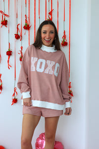 Woman wearing a pink 'KOKO' sweatshirt and shorts in front of a decorative wall with red ribbons and strawberries.
