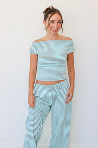 A woman modeling a matching two-piece loungewear set featuring a light blue and white striped off-the-shoulder crop top and ruffle wide leg pants, standing against a plain white wall.