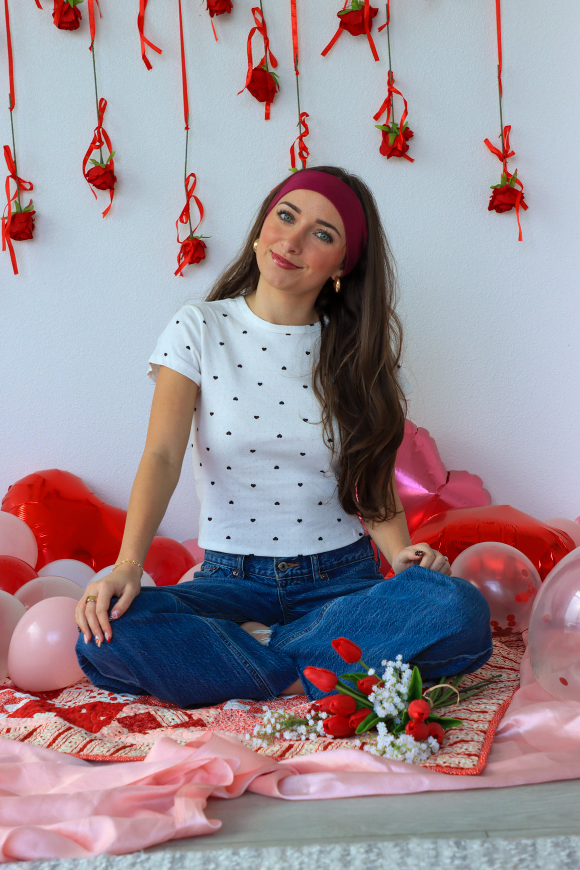 Woman sitting on a decorated floor with balloons and flowers, wearing a white polka dot shirt and blue jeans.