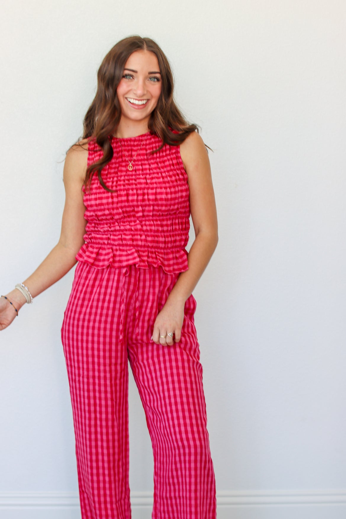 A woman modeling a sleeveless red and pink gingham tank and pant set with a smocked waist and ruffle detail, standing against a plain white wall.