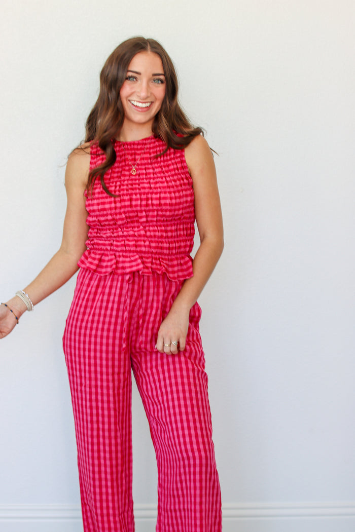 A woman modeling a sleeveless red and pink gingham tank and pant set with a smocked waist and ruffle detail, standing against a plain white wall.