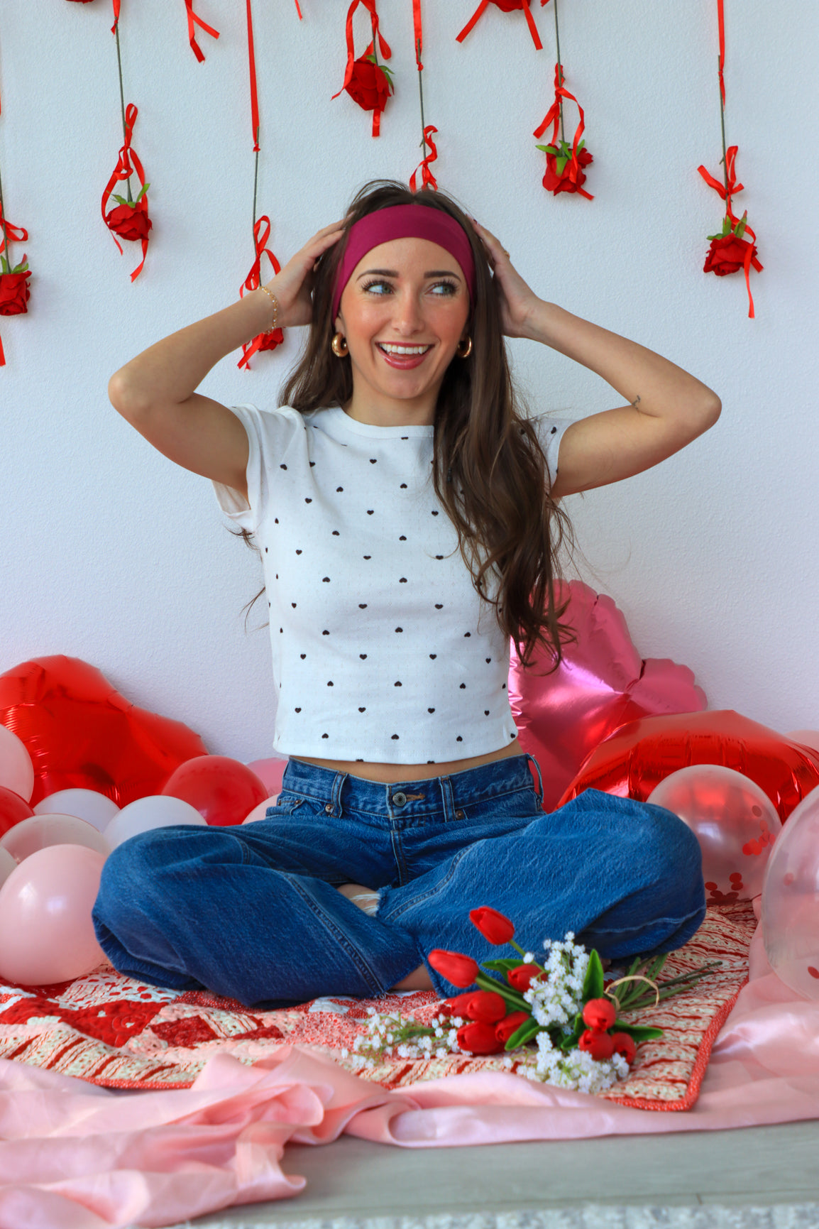 Woman sitting on a bed with balloons and flowers, wearing a white polka dot top and blue jeans.