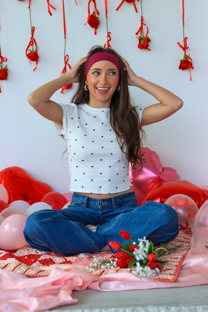 Woman sitting on a bed with balloons and flowers, wearing a white polka dot top and blue jeans.