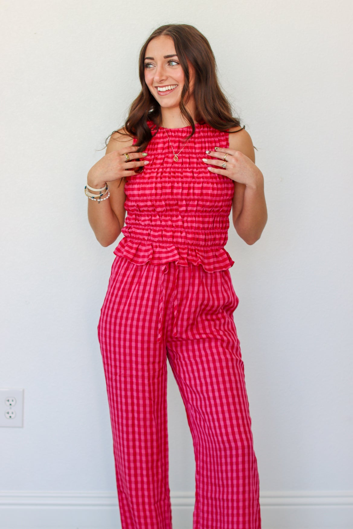 A woman modeling a sleeveless red and pink gingham tank and pant set with a smocked waist and ruffle detail, standing against a plain white wall.