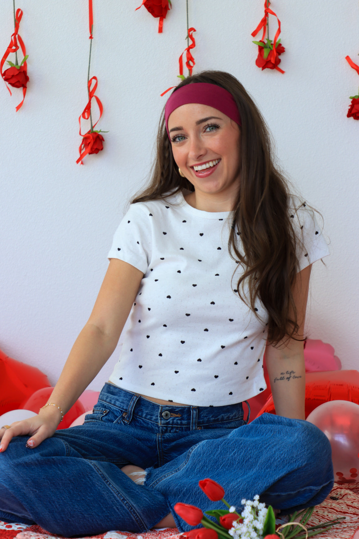 Woman wearing a white polka dot shirt and blue jeans sitting on a floor with red decorations and balloons.