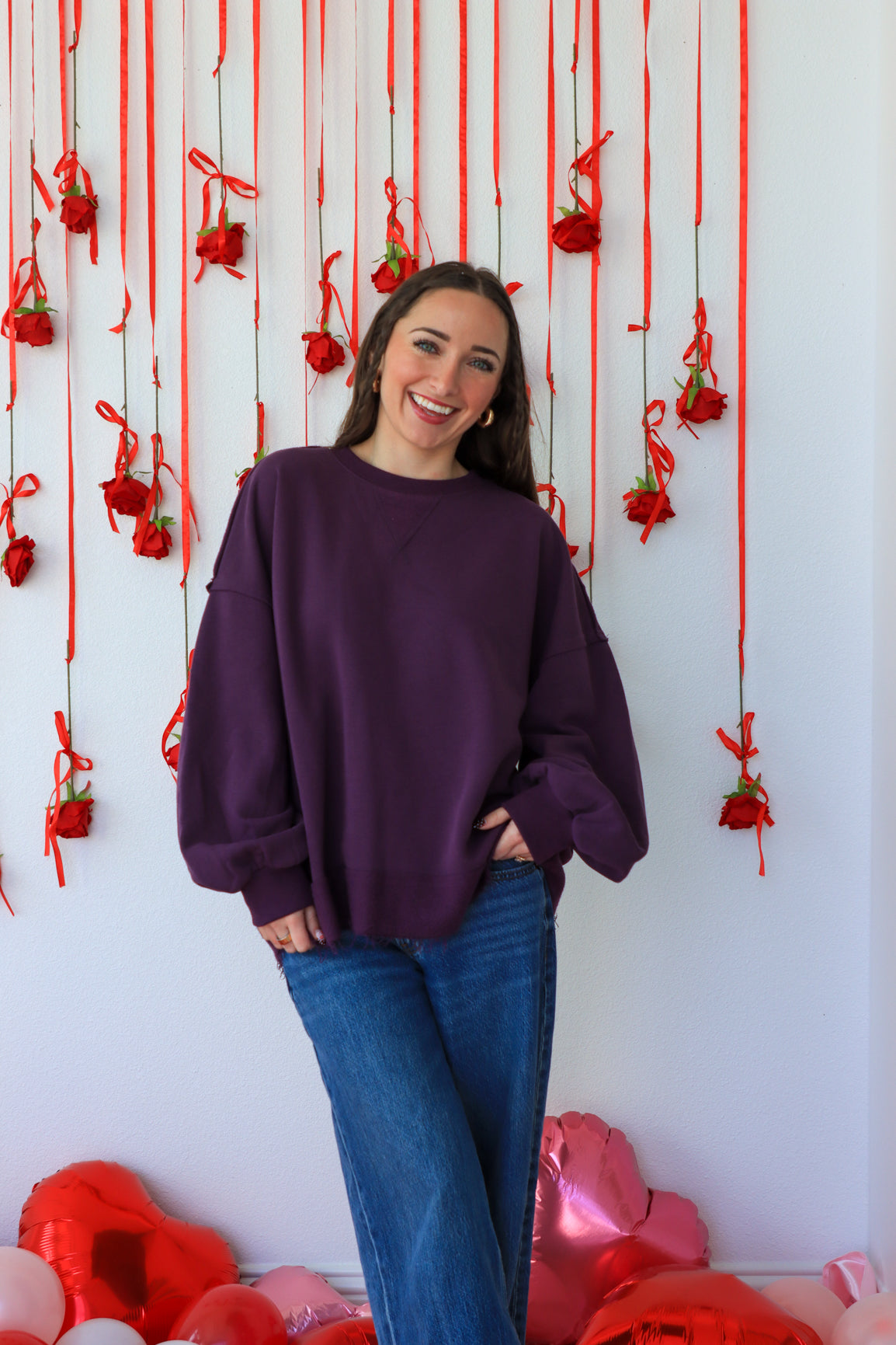 Woman in a purple sweater and blue jeans standing in front of a decorative wall with red ribbons and heart-shaped balloons.