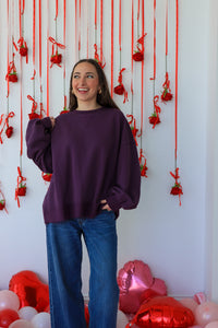 Person wearing a purple sweatshirt and blue jeans standing in front of a wall with red ribbons and balloons.