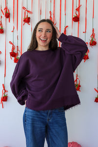 Woman wearing a purple sweater and blue jeans standing against a decorative wall with red ribbons and flowers.