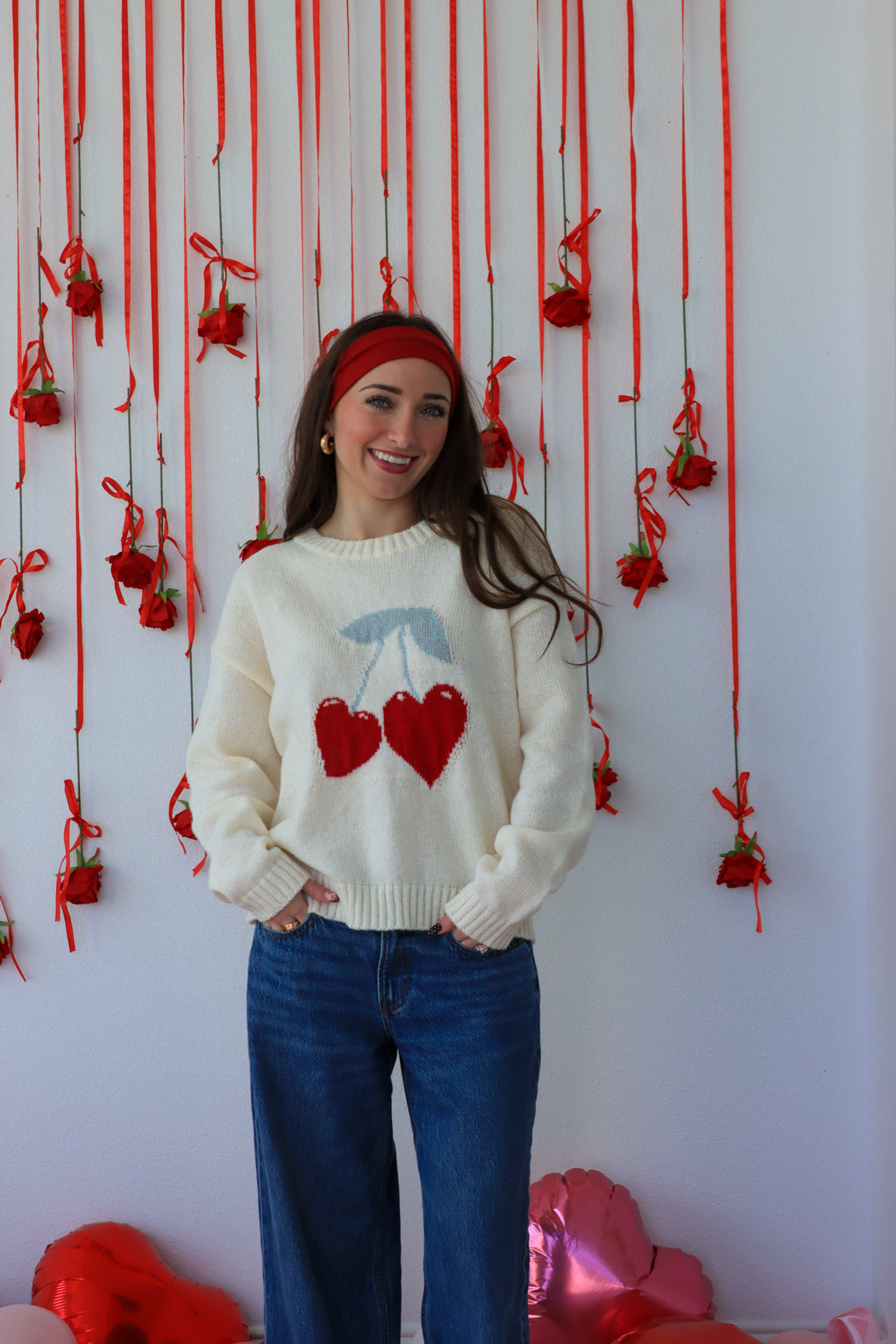 Woman wearing a white sweater with red heart design in front of a decorative wall with red ribbons and heart-shaped decorations.