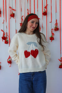 Woman wearing a cream sweater with red heart design in front of a decorative wall with red ribbons and flowers.