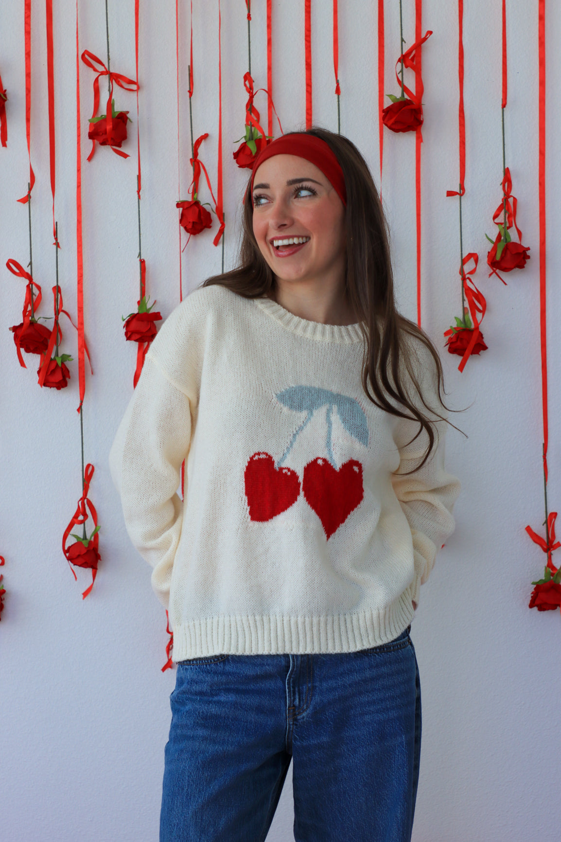 Woman wearing a sweater with cherry design in front of a decorative wall with red ribbons and flowers.