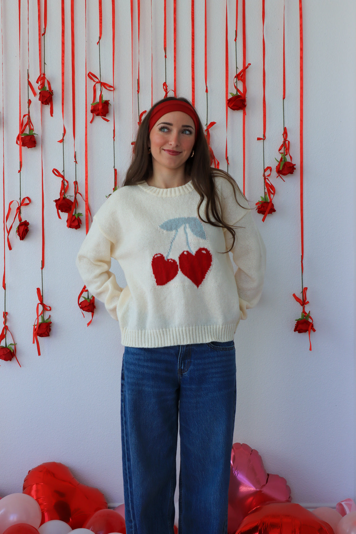 Woman wearing a sweater with heart designs in front of a decorative wall with red ribbons and balloons.