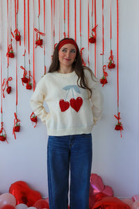 Woman wearing a sweater with heart designs in front of a decorative wall with red ribbons and balloons.