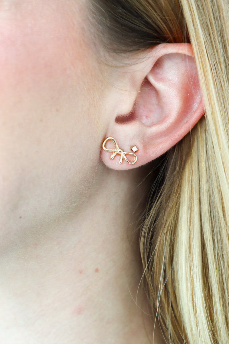 Gold bow-shaped earring worn by a person with blonde hair.