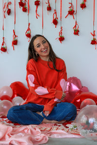 Woman in a red sweater with pink bunny ears surrounded by balloons and decorations on a white wall.