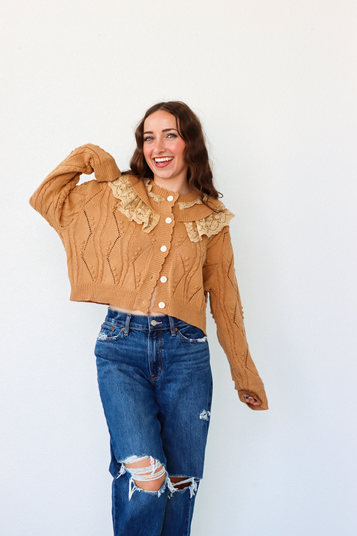 Woman wearing a brown cable knit sweater with lace details and blue jeans on a white background