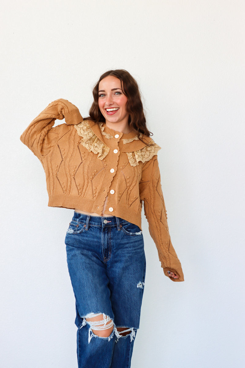 Woman wearing a brown cable knit sweater with lace details and blue jeans on a white background