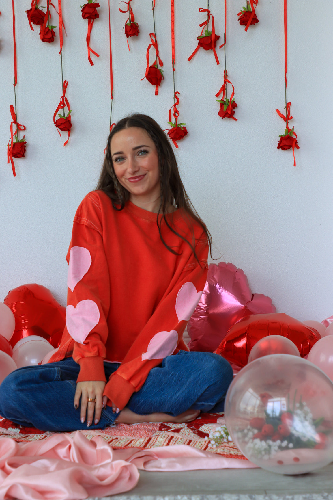 Woman in a red sweater with heart patterns sitting among pink balloons and decorations on a white wall.