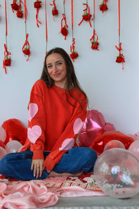 Woman in a red sweater with heart patterns sitting among pink balloons and decorations on a white wall.
