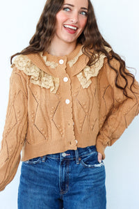 Woman wearing a beige cable knit cardigan with lace details and blue jeans on a light background