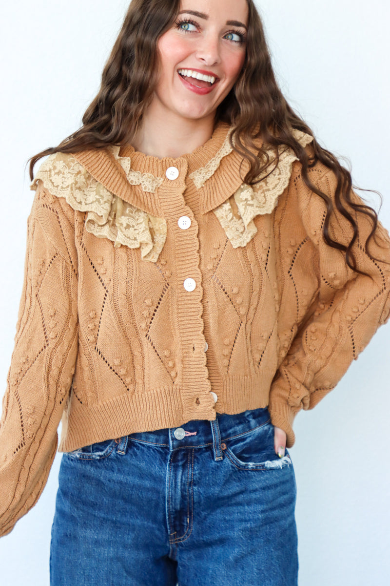 Woman wearing a beige cable knit cardigan with lace details and blue jeans on a light background
