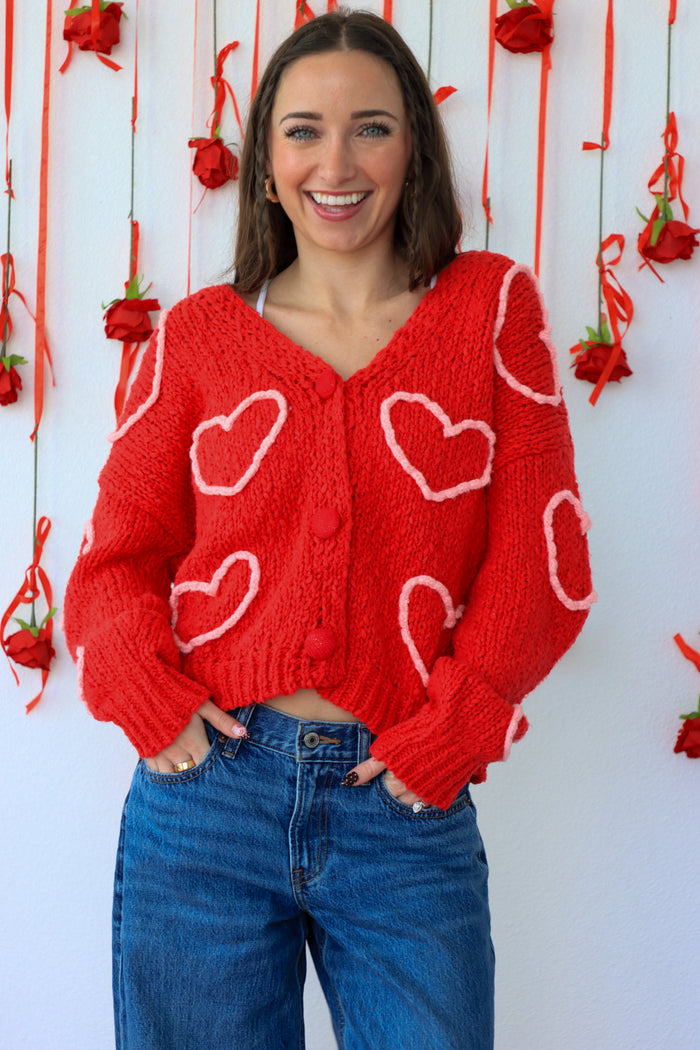 Woman wearing a red cardigan with heart patterns against a decorative background.