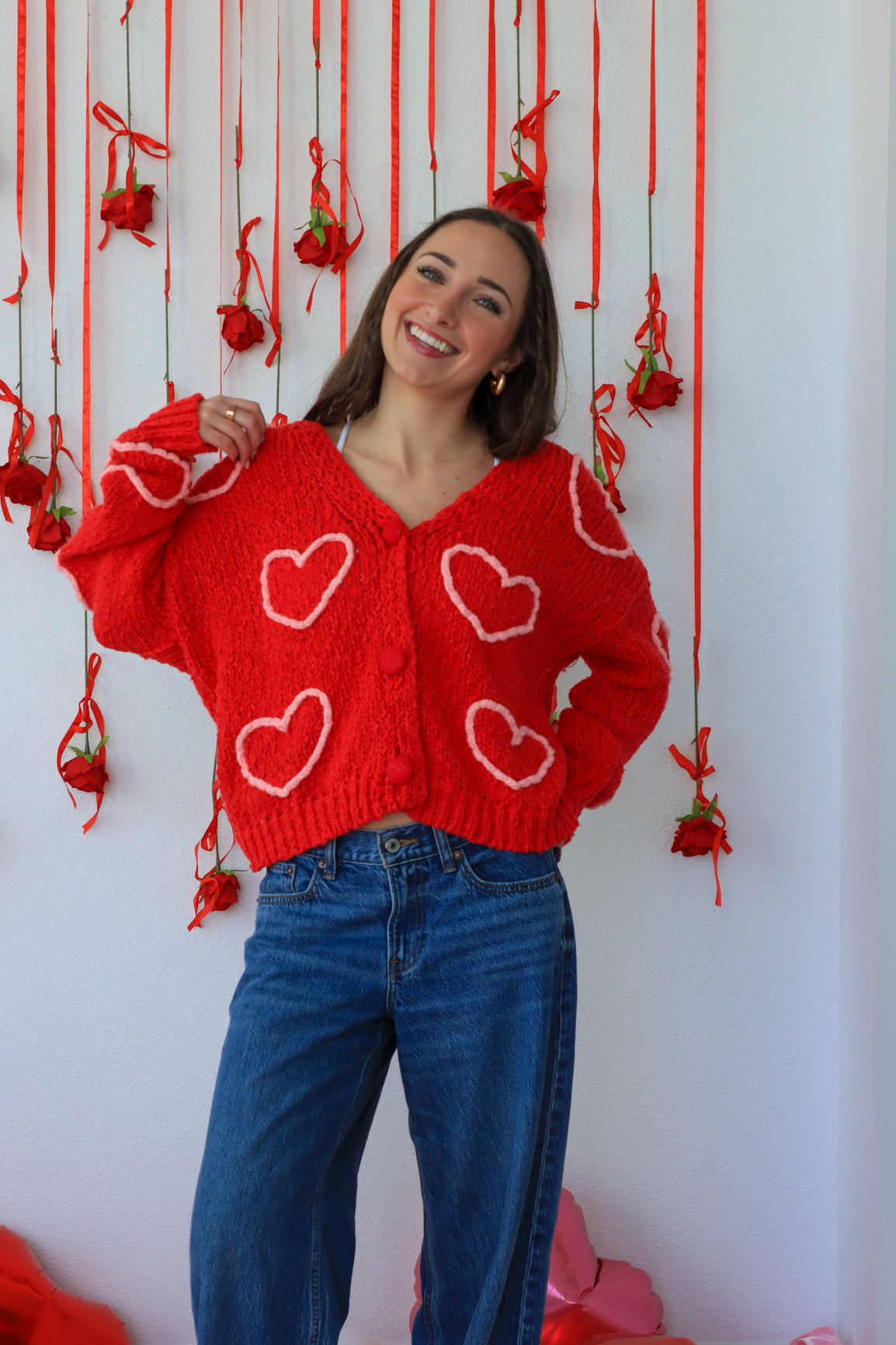 Woman wearing a red cardigan with heart patterns in a decorative setting with red ribbons and small red bags.