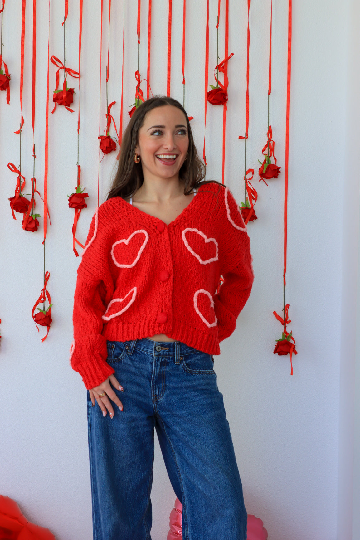 Woman wearing a red sweater with heart patterns in front of a decorative wall with red ribbons and small red bags.