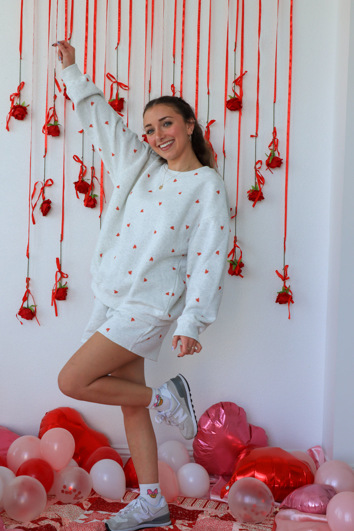 Woman in a light blue sweatshirt with red polka dots and white shorts posing in front of a wall decorated with red ribbons and balloons.