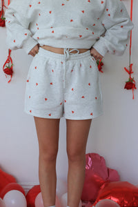 Person wearing a light gray sweatshirt and shorts with red heart patterns, standing against a white wall with red balloons.