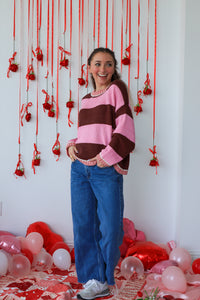 Woman in a striped sweater and blue jeans standing in front of a wall with red decorations and balloons on the floor.