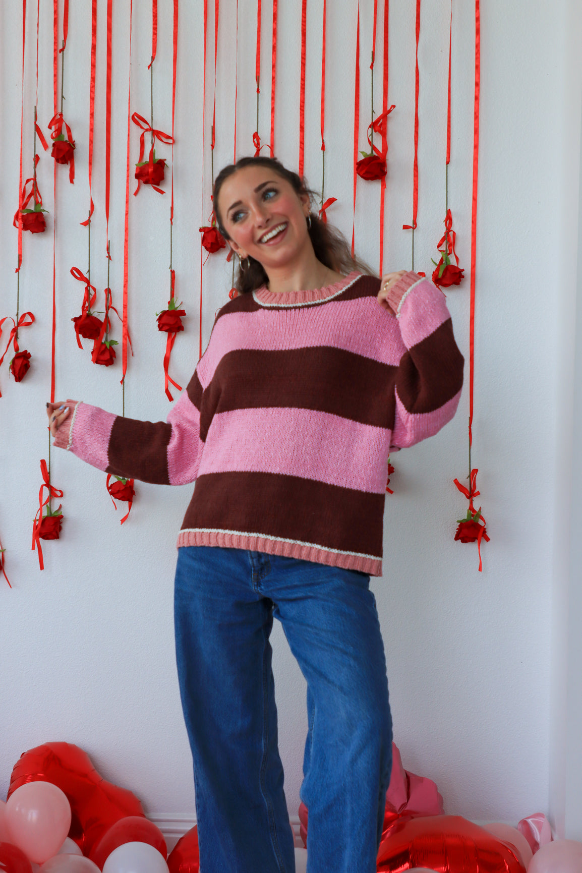 Woman wearing a pink and brown striped sweater with red decorations on a white wall.