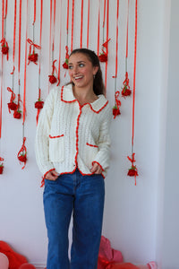 Woman wearing a white cardigan with red trim standing in front of a decorative wall with red ribbons and flowers.