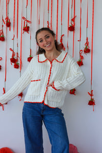 Woman wearing a white knitted cardigan with red trim against a decorative background.