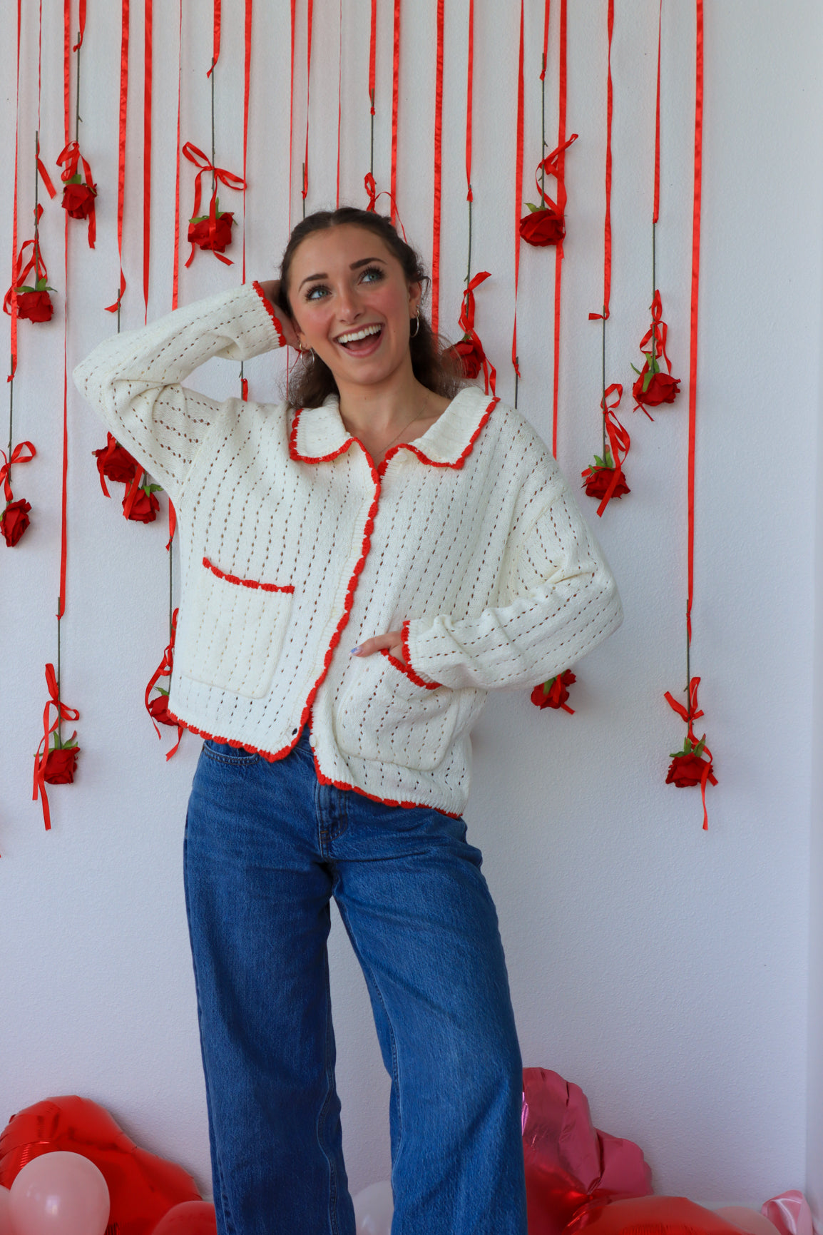Woman wearing a white knitted cardigan with red trim against a decorative wall with red ribbons and balloons.