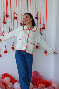 Woman wearing a white cardigan with red trim standing in front of a decorative wall with red ribbons and balloons.