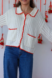 Person wearing a white crocheted cardigan with red trim against a white background