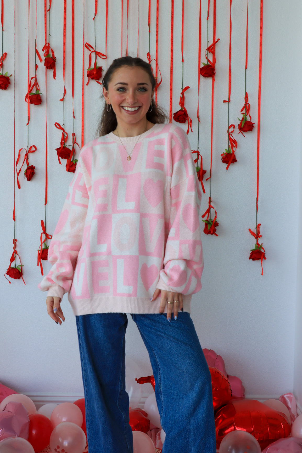 Woman wearing a pink and white patterned sweater with blue jeans standing in front of a decorative wall with red ribbons and balloons.