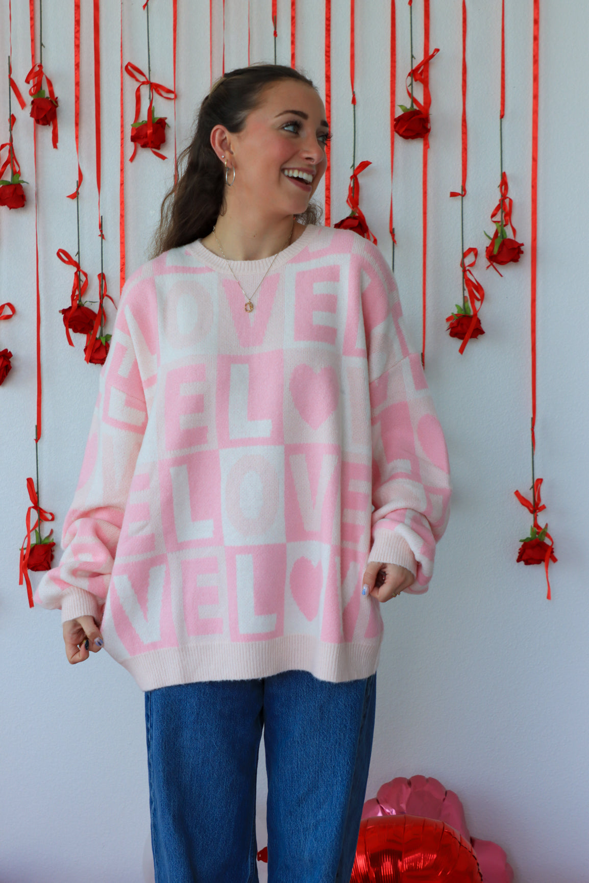 Person wearing a pink sweater with text, standing against a decorative wall with red ribbons and heart shapes.