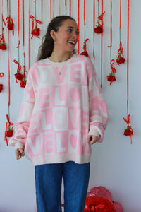 Person wearing a pink sweater with text, standing against a decorative wall with red ribbons and heart shapes.