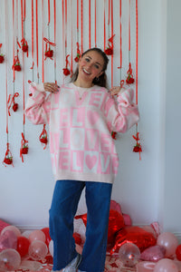 Woman wearing a pink sweater with text and blue pants standing in front of a decorative wall with red ribbons and balloons.