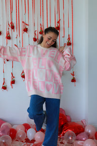 Woman wearing a pink sweater with 'LOVE' printed on it, standing in front of a decorative wall with red ribbons and balloons.