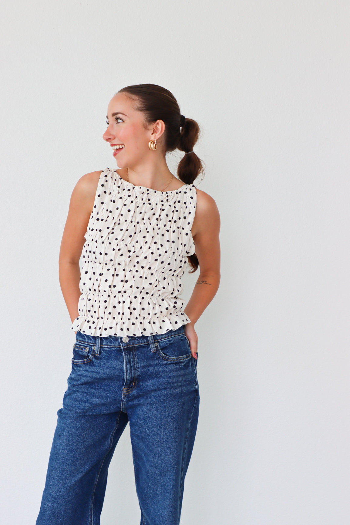 Woman wearing a white sleeveless top with black polka dots and blue jeans against a white background