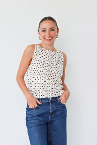 Woman wearing a white sleeveless top with black polka dots and blue jeans against a white background