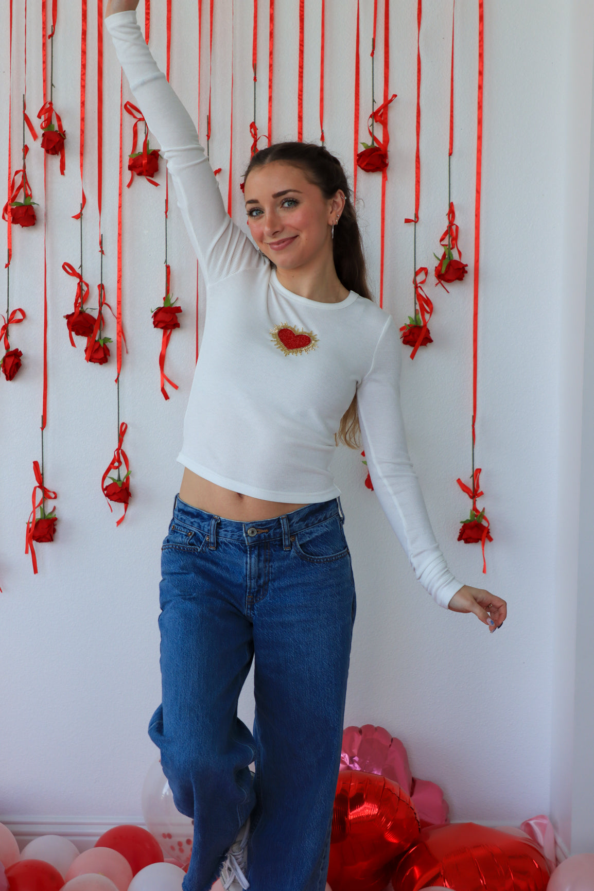 Person wearing a white long-sleeve top with red heart designs and blue jeans, standing against a wall with red decorations.