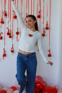 Person wearing a white long-sleeve top with red heart designs and blue jeans, standing against a wall with red decorations.
